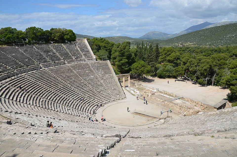 A vast semicircle of grey stone seating rows climbing a wooded hillside, looking down on a round orchestra and the low ruins of a stage building, with pine forest and mountains beyond.