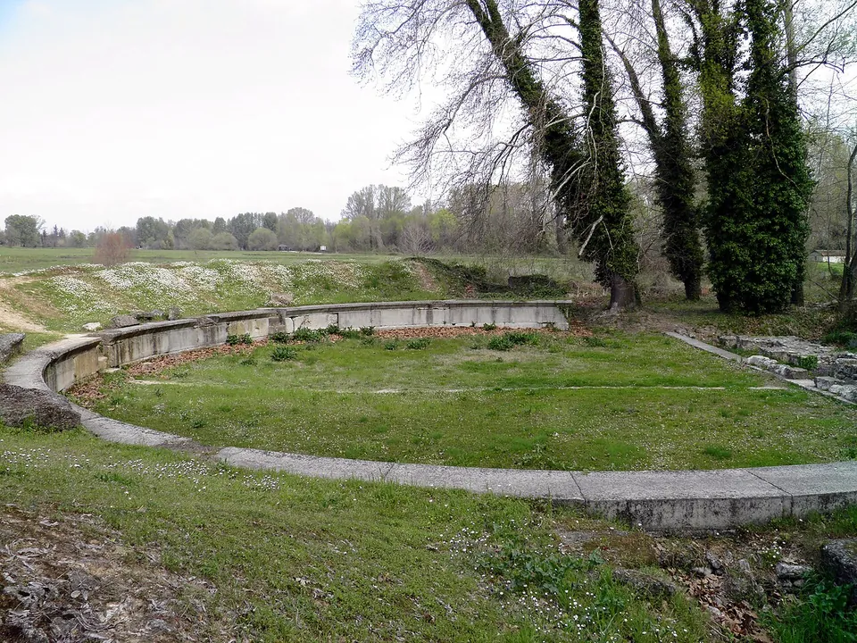 A grassy horseshoe-shaped space ringed by a low curved wall of stone, with bare trees and ivy-covered trunks on the right and flat fields beyond.