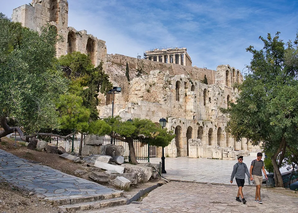 The tall outer wall of a ruined theatre built of stone, pierced by rows of arched openings, seen from a paved path among trees, with a temple's columns on the rock above it.