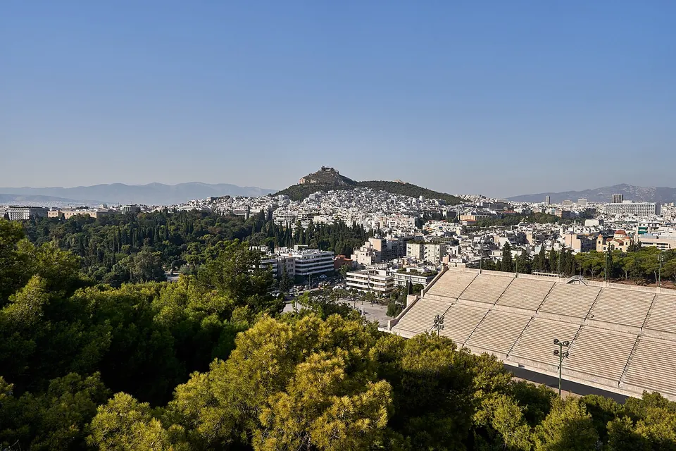 Looking down from a pine-covered hill onto the marble tiers of a stadium at the lower right, with the white city of Athens spreading behind towards a single rocky peak.