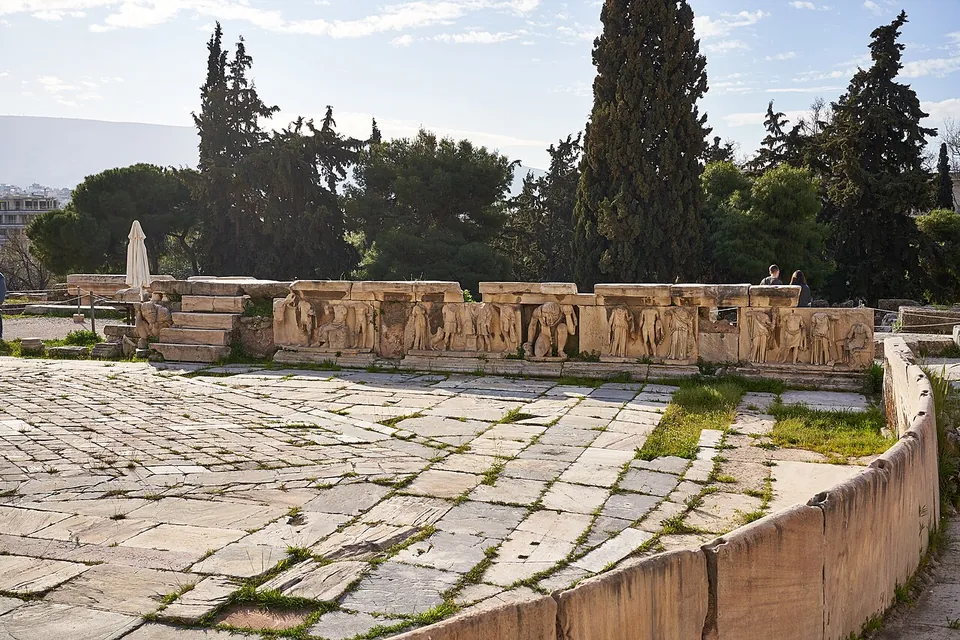 A low marble wall carved with a frieze of figures in relief, some crouching, at the edge of a floor of worn stone slabs, with cypresses and pines behind.
