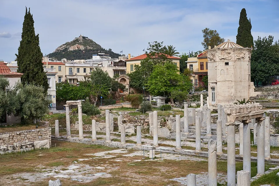 A broad open courtyard of bare ground and worn paving with rows of broken marble column shafts, an octagonal marble tower on the right, tall cypresses and houses behind, and a steep wooded hill in the distance.