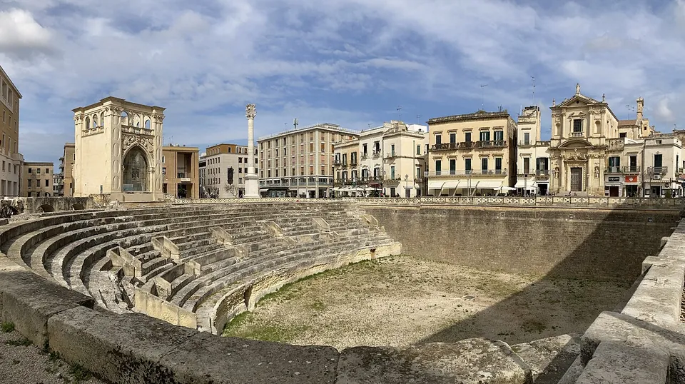 Curving tiers of stone seating and a grassy arena floor lie below a square ringed by pale stone palaces, an ornate Baroque facade and a column topped by a statue, under a cloudy sky.