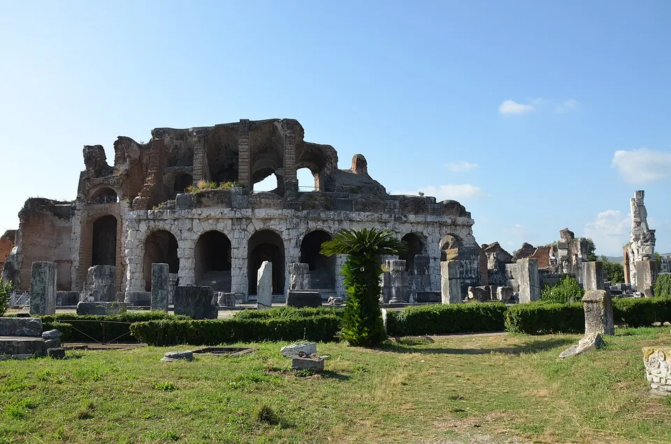 A long run of ruined arcading in pale stone, with a higher, broken storey of brick piers behind it, seen across a lawn with clipped hedges, a palm and scattered stone fragments.