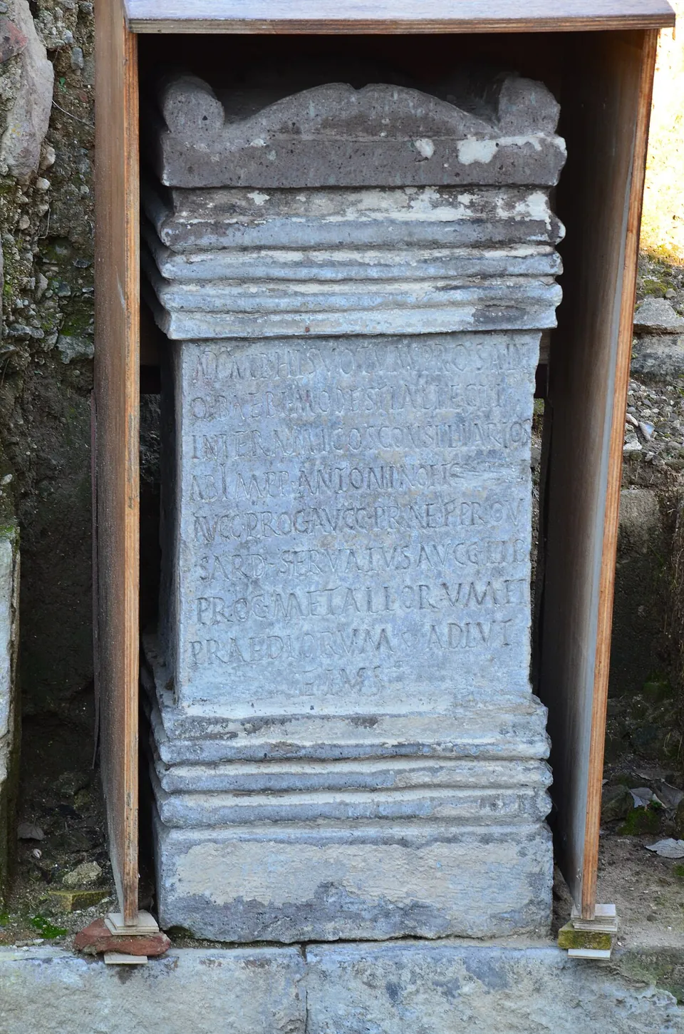 A stone altar with a moulded base and crown, its face cut with nine lines of Latin capitals, standing in an open wooden case against an excavated wall.