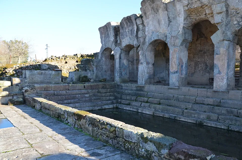 A row of round arches on heavy pillars of grey volcanic stone, the top of the wall broken and uneven, rising above stone steps that run down into a rectangular pool of still water.