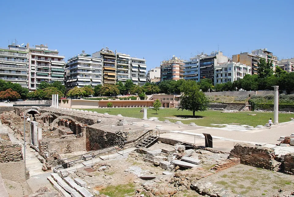 A broad excavated square of grass and paving stones, with a single tall column standing on the right, low stone walls and vaulted openings in the foreground, and apartment blocks lining the far side.