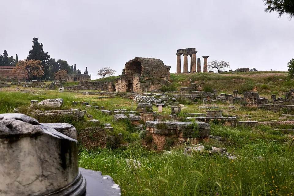 A wide grassy field of excavated walls, column bases and fragments, with the brick ruin of a vaulted building in the middle distance and seven Doric columns of a temple on a rise beyond.