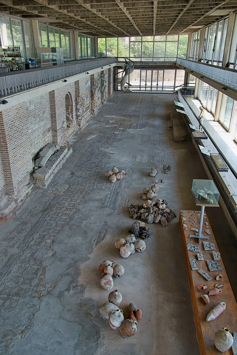A long modern hall with glass walls and a gallery seen from above, its floor a large worn mosaic of geometric patterns, with amphorae laid out on it, a display case and a brick wall with arches along one side.