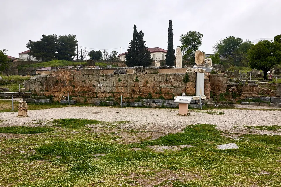 A long platform of squared stone blocks across a gravel square, with a few carved fragments set on top of it, an information board in front, and cypresses and village houses behind.