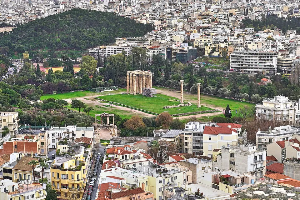 Looking down over red roofs and apartment blocks to a green precinct where a cluster of tall columns and two lone ones stand on a lawn, with an arched gateway at its corner and a wooded hill beyond.