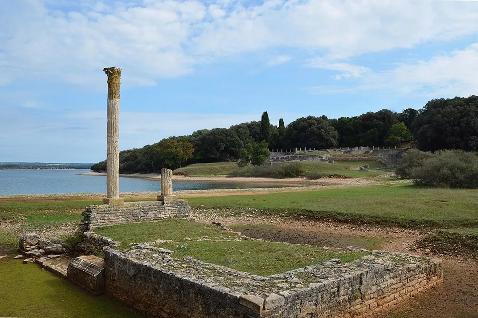 A tall stone column with a worn capital stands at the corner of a low rectangular stone platform on grass, a shorter column stump behind it, with the bay and a wooded shore beyond.