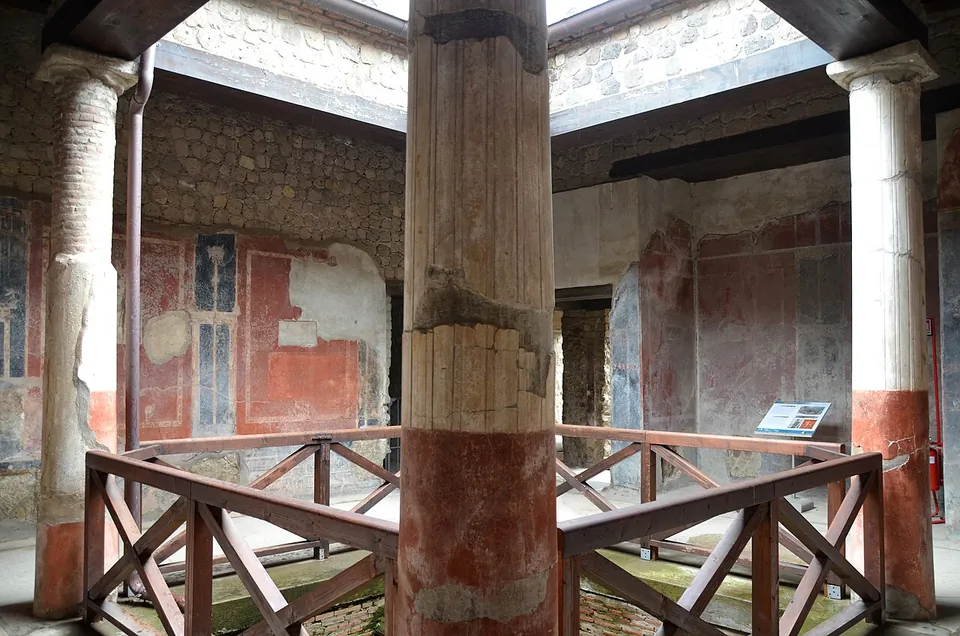 A small courtyard of four plastered columns, red at the base, around a sunken basin fenced with a wooden rail, the walls behind painted red and black, under an opening in a modern roof.