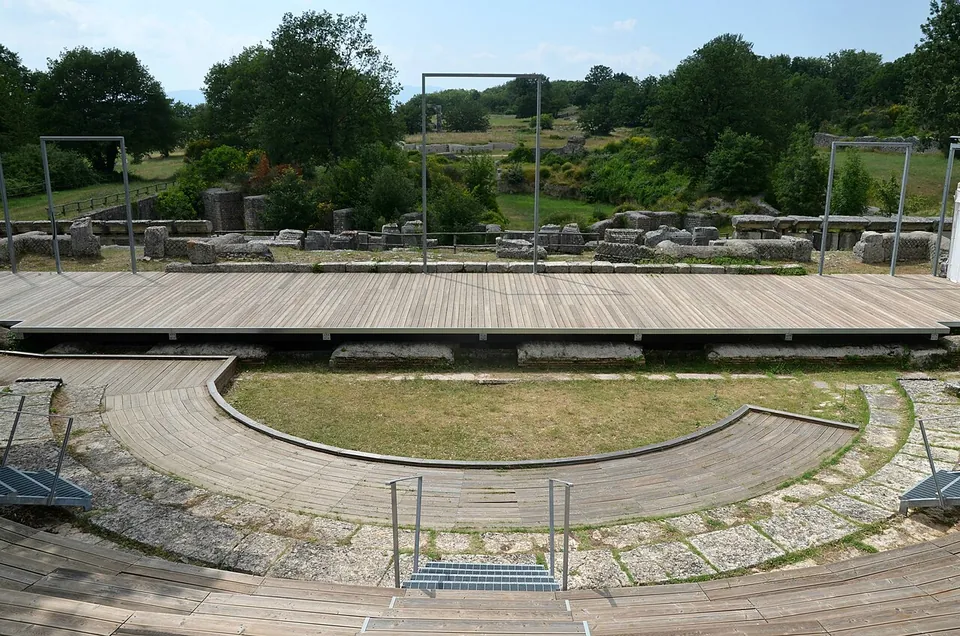 The semicircle of the theatre seen from the seating, fitted with modern timber benches and a timber stage, with steel frames on the stage and the ruins of the town beyond.