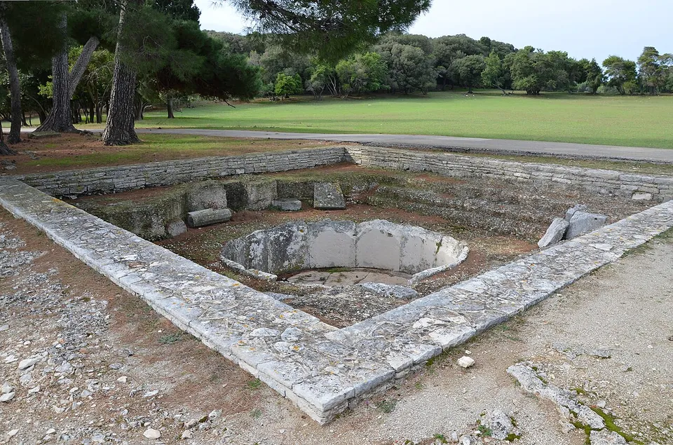 A square sunken room edged with large stone slabs, with a round stone basin set into its floor and a lawn and pines beyond.