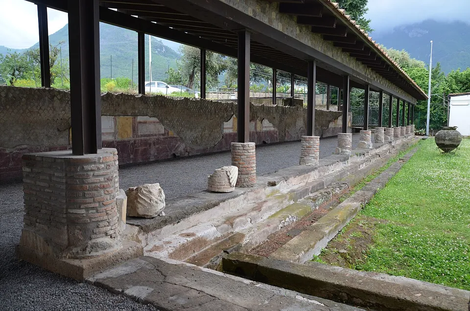 A long covered portico on brick-built column stumps, with fragments of carved stone lying beside a stone-edged channel and a strip of lawn, and wooded mountains rising behind under cloud.