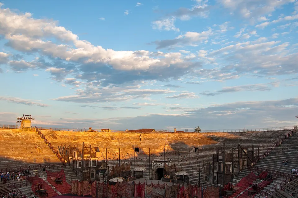 Stone seating tiers lit by low evening sun under a cloudy sky, with a large stage set of towers and painted scenery built across one end of the arena floor and spotlight rigs at the top of the seating.