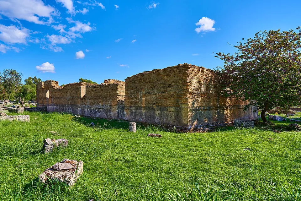 A long rectangular building of reddish brick and stone, its walls standing well above head height, on a green lawn under a blue sky.