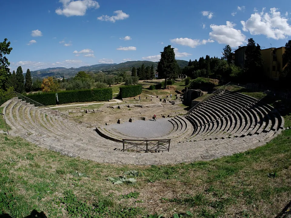 The curved tiers of the theatre seen from the top of the seating, with a railing round the orchestra, hedges and cypresses behind the stage area and a wide view of green hills under a blue sky.