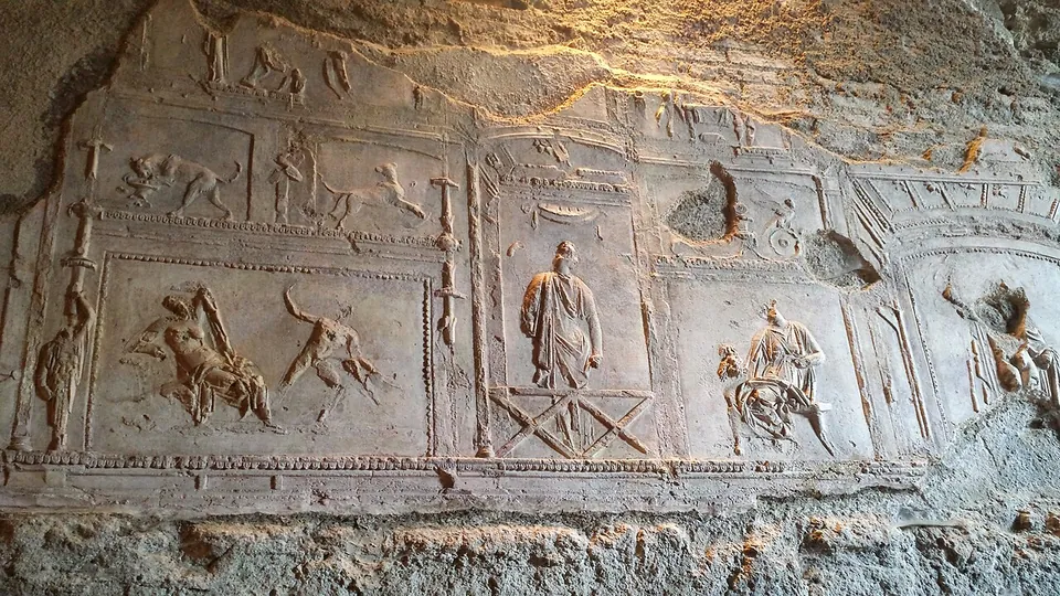 A frieze of low-relief panels in pale stucco on a rough wall, framed by raised borders and lit from one side. Small human figures and animals fill the panels, and a standing draped figure is at the centre.