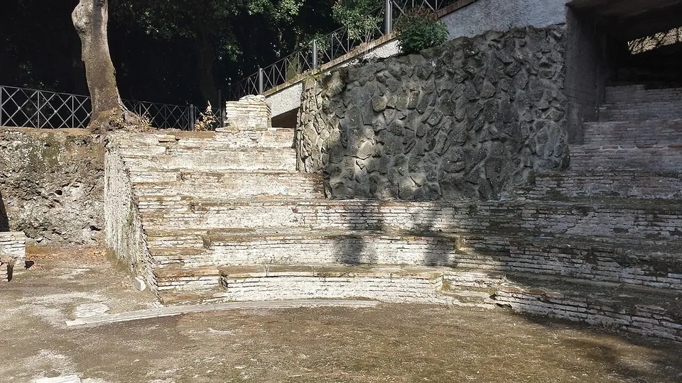 Curved tiers of stone and brick seating rising from a flat floor, with a wall of irregular dark stones beside them. A modern concrete walkway and railing run along the top, among trees.