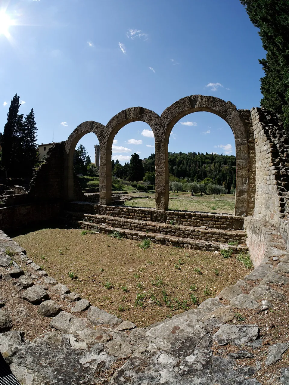 Three round stone arches on square piers, seen across a sunken stone-edged basin with steps along its far side, under a bright sky.