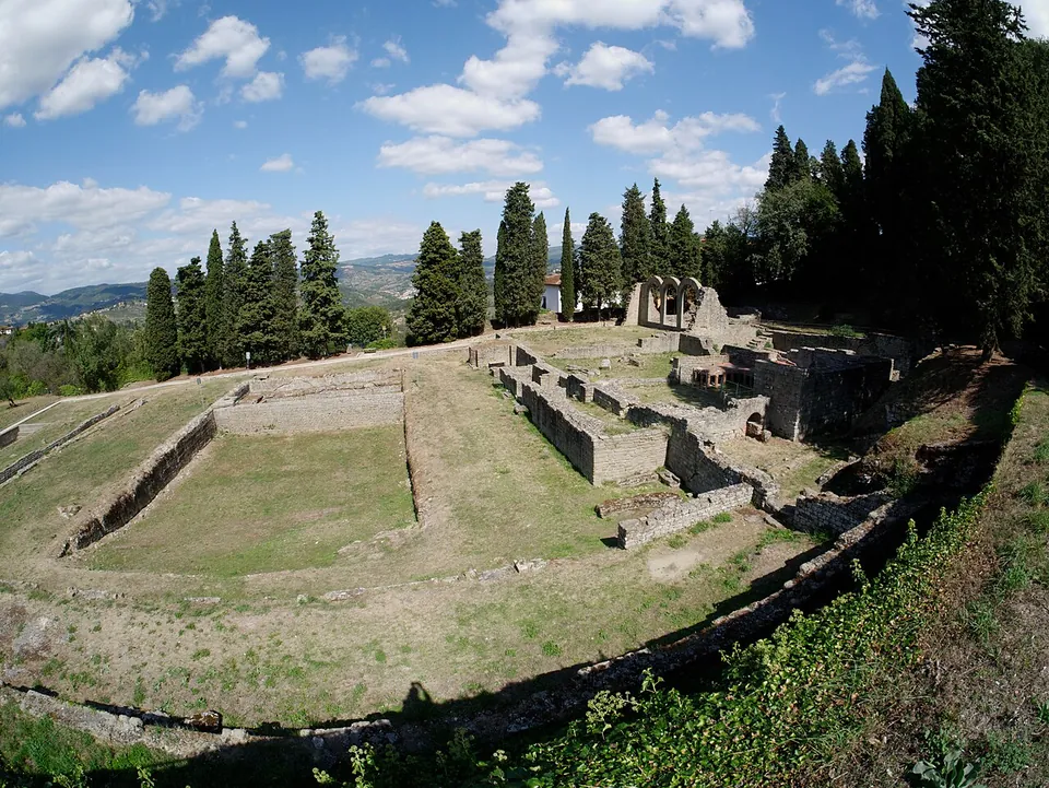 A wide view across a grassy terrace with two large rectangular sunken basins edged in stone, the walls of the bath rooms beyond them and the three arches in the distance, with cypresses and green hills on the horizon.