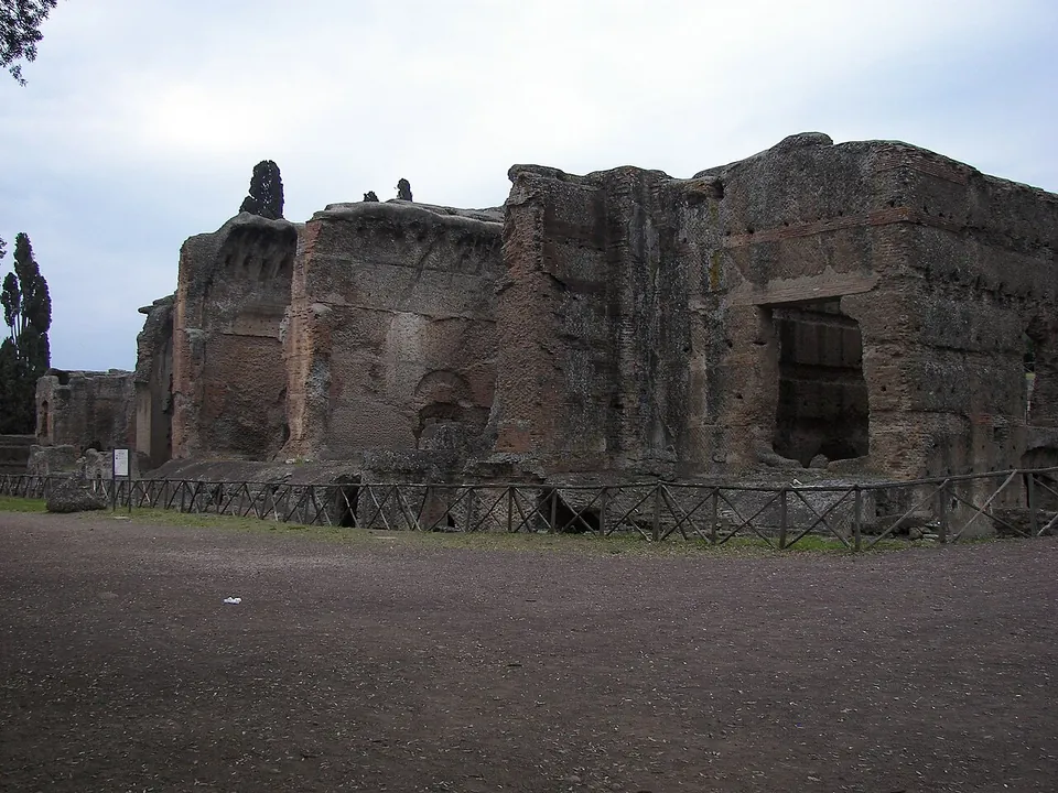 Massive ruined walls of brick-faced concrete several storeys high, with the springing of vaults and a large rectangular opening, behind a low wooden fence across a bare gravel space.