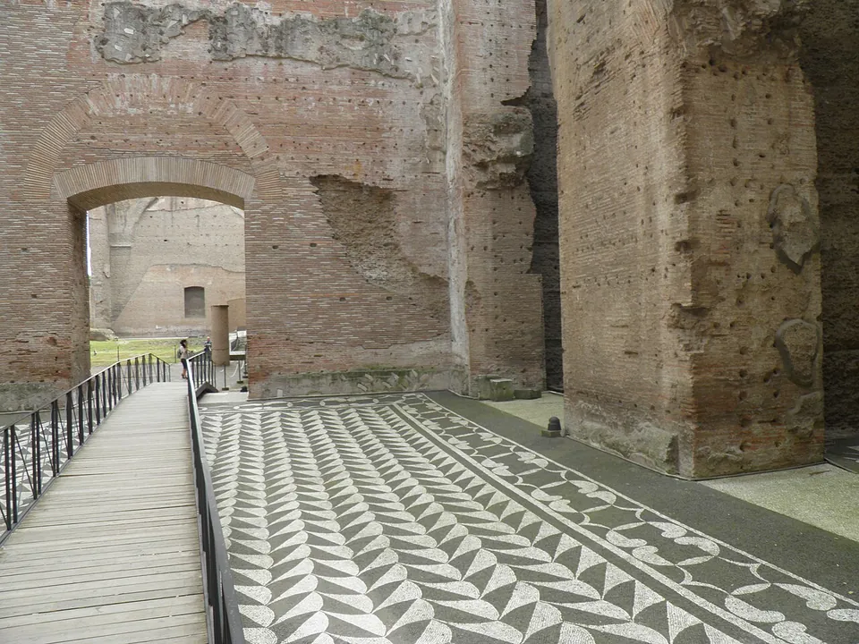 A floor mosaic of black and white geometric pattern beside a raised wooden walkway with railings, between high brick walls with an arched doorway.