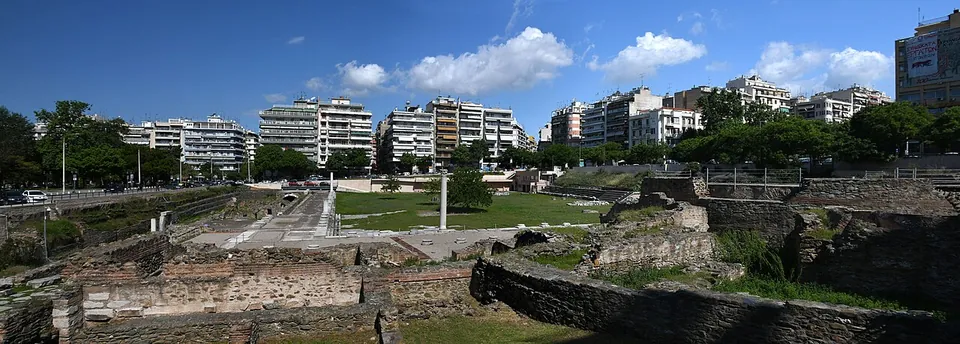A panorama of the excavated forum under a blue sky, with walls of brick and stone in the foreground, the grassed square and a tall column beyond, and the city's apartment blocks all around.