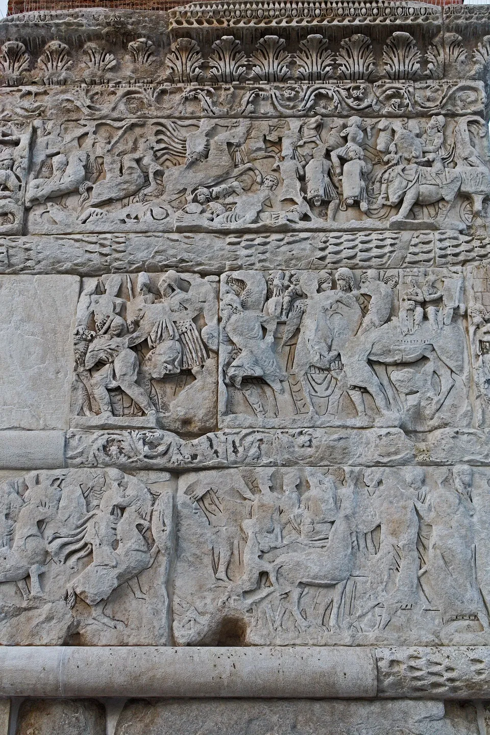 Weathered marble relief in stacked bands, crowded with horsemen, soldiers and figures in long robes, separated by carved mouldings of leaves and garlands.