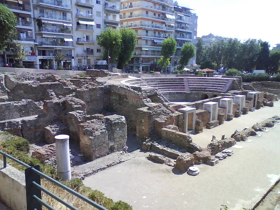 A small semicircle of stone seating rows behind ruined walls of stone and brick, with a row of square stone bases along a paved path and apartment blocks behind.