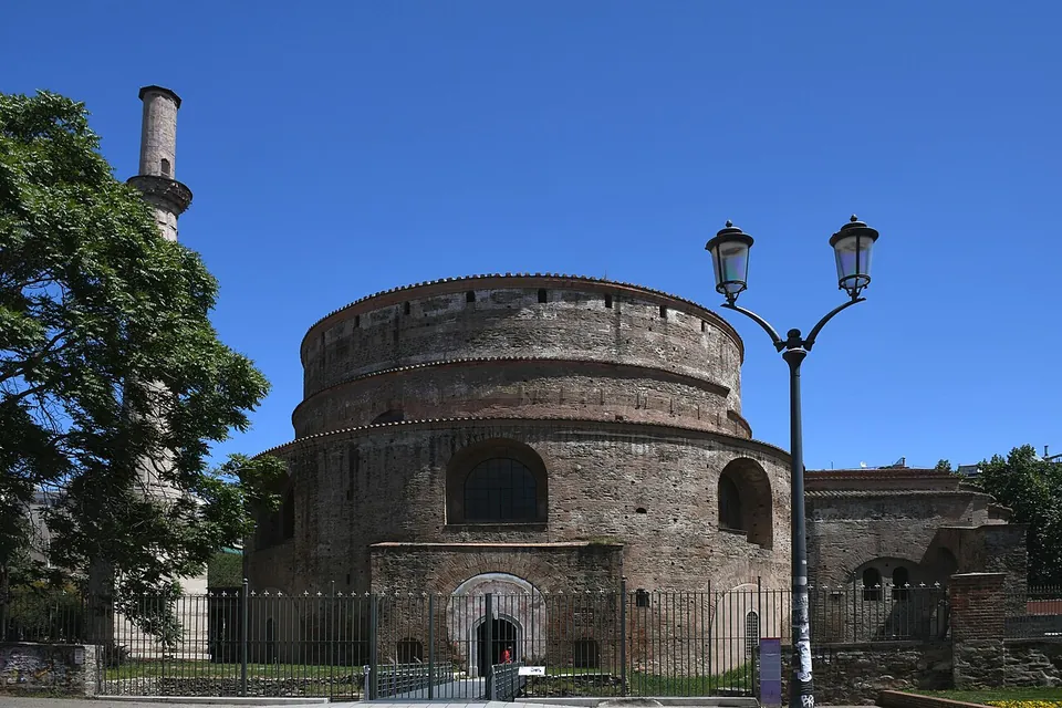 The round brick building behind a black railing with a gate, a minaret rising on the left behind a tree, and a street lamp on the right.