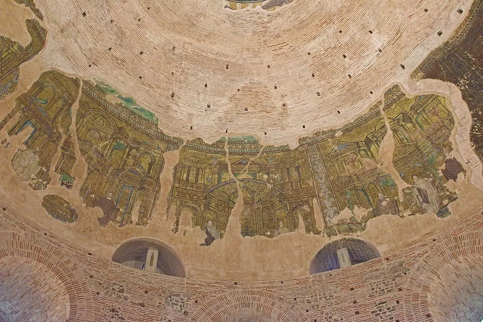 The inside of a brick dome, ringed by a band of mosaic in gold, green and brown showing rows of fantastic buildings with columns and arches, broken by bare patches, above two arched windows.