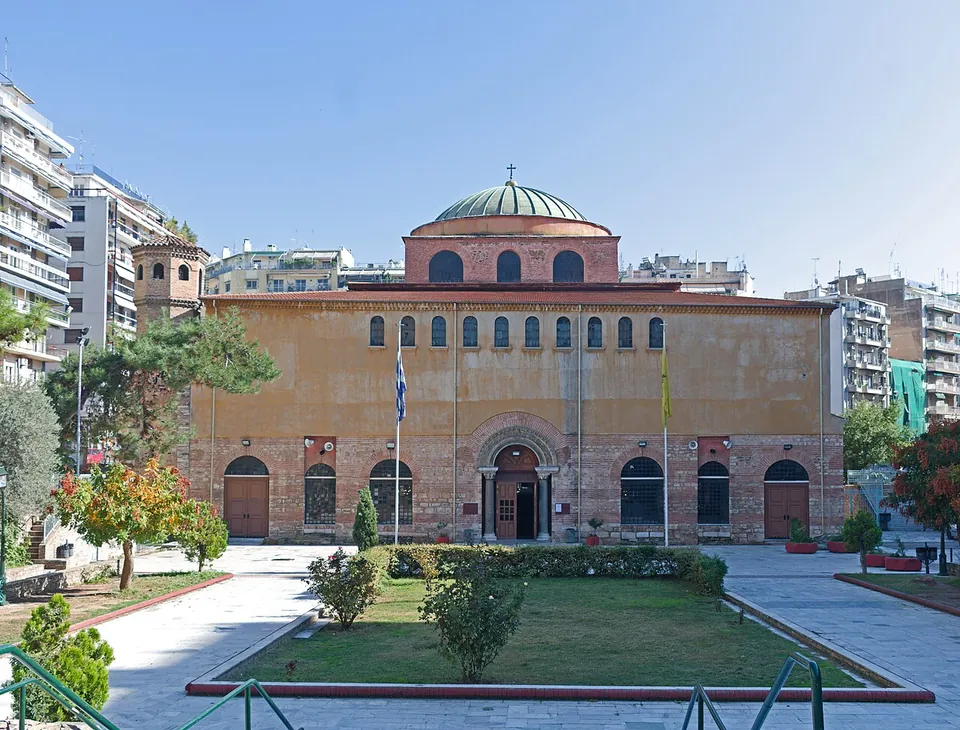 The broad west front of a church in ochre plaster and brick under a green-roofed dome, with arched doorways and windows, a garden of shrubs and a paved square in front and apartment blocks around.