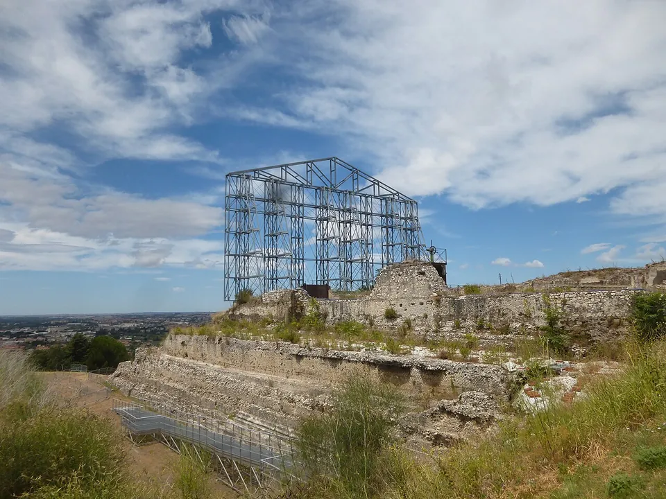 Grassy terraces of rough stone walls step down towards a wide plain on the horizon, with a tall open steel framework standing on the ruins at the top.