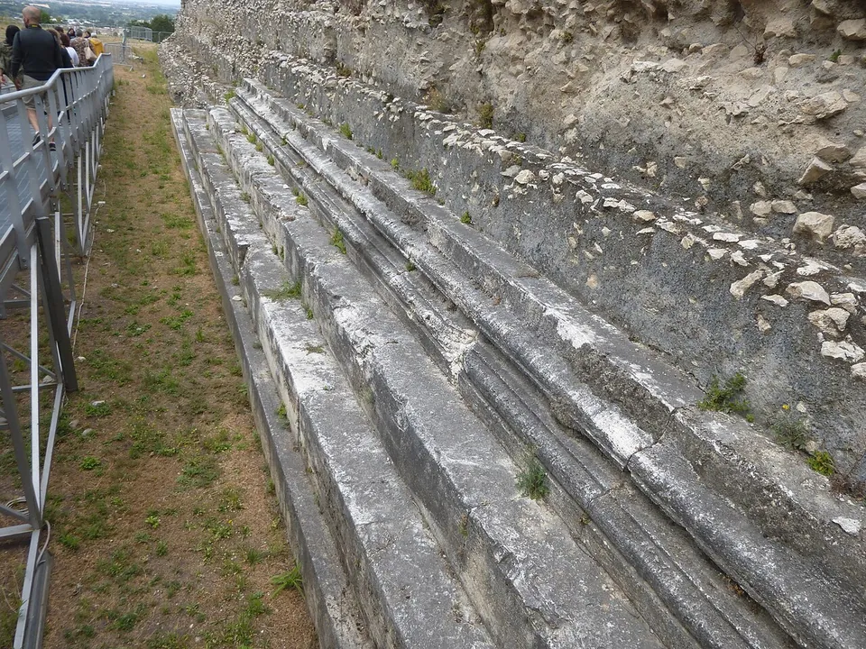 Long courses of moulded grey stone run along the base of a rubble wall, beside a strip of grass and a metal railing.