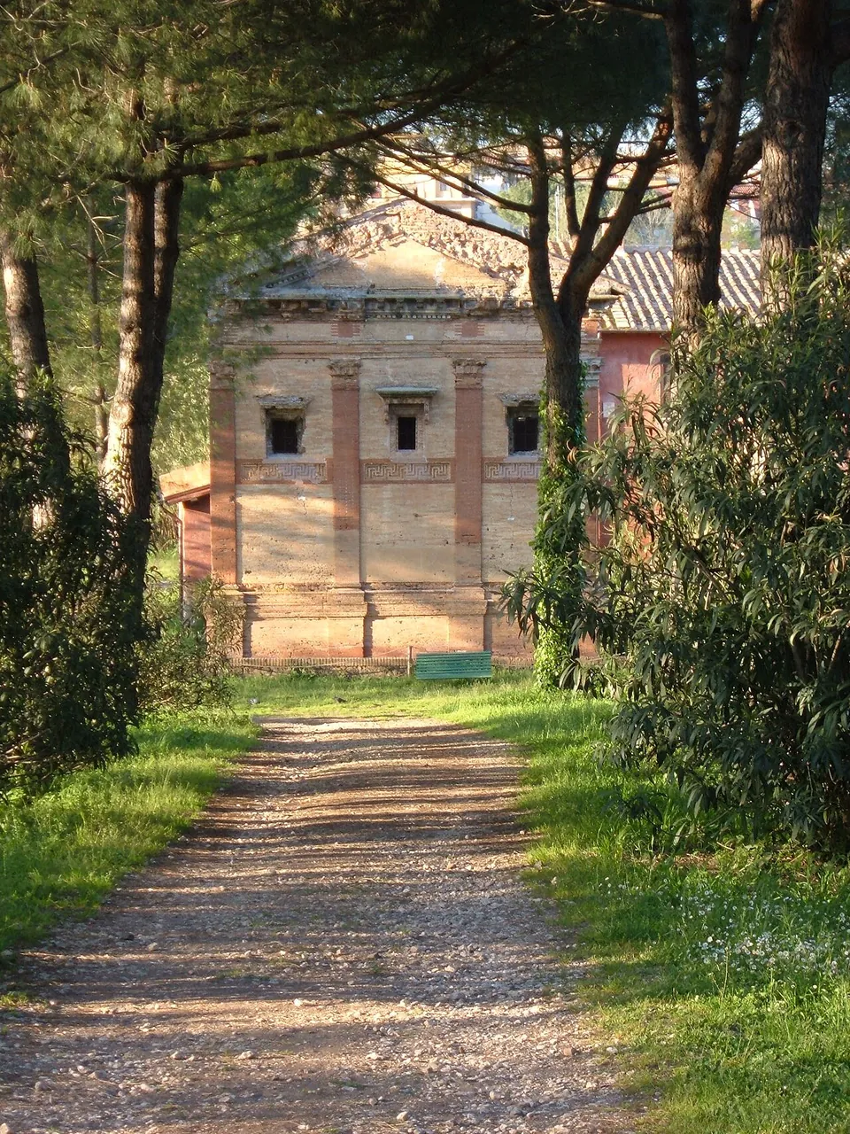 A gravel track between grass verges and pine trees leading straight to the brick front of the tomb at its far end.