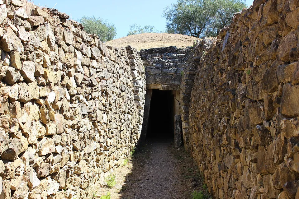 A corridor walled in dry-laid stone leads to a dark doorway framed by stone slabs, with the grassy curve of a burial mound above it.