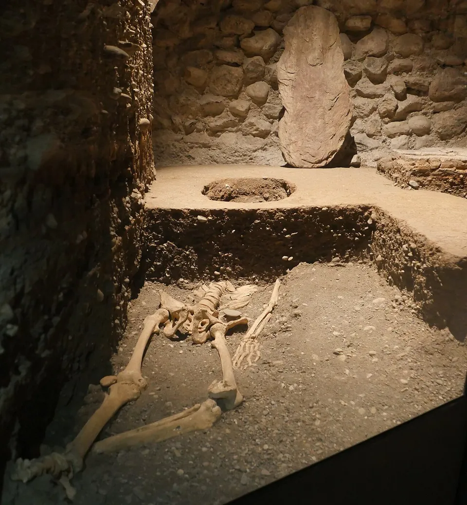 A skeleton lying in an excavated grave cut into a layered earth floor, in a dim underground space with a rough stone wall and a tall standing stone behind.