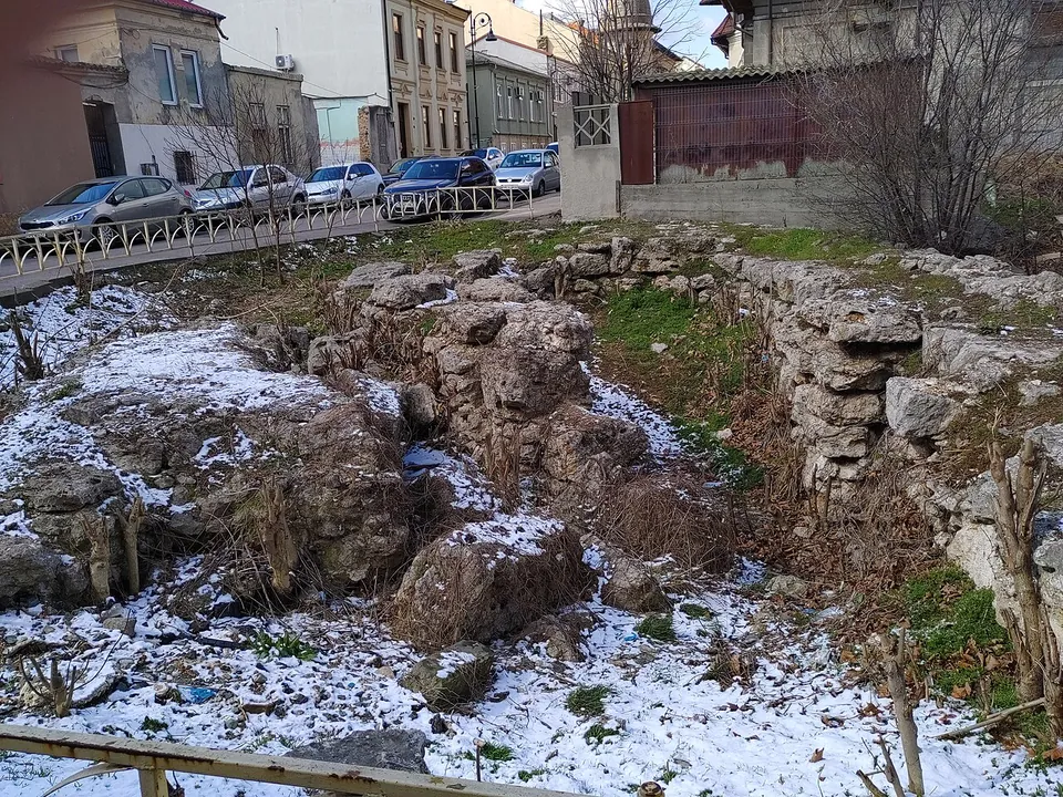 Rough blocks and rubble of a ruin in a sunken plot dusted with snow, behind a railing on a city street lined with parked cars and old houses.