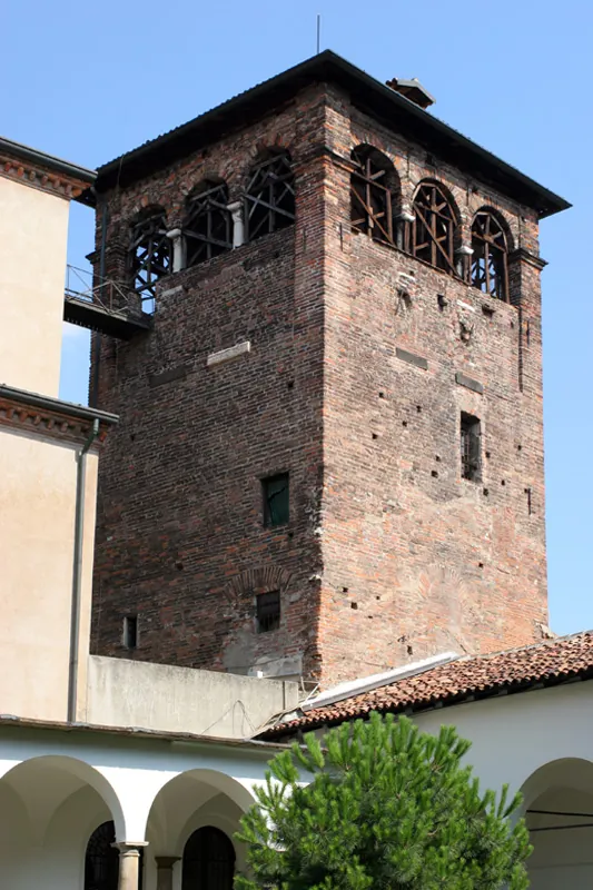 A tall square brick tower with a row of arched openings below its tiled roof, rising behind the arcade and roofs of a cloister.
