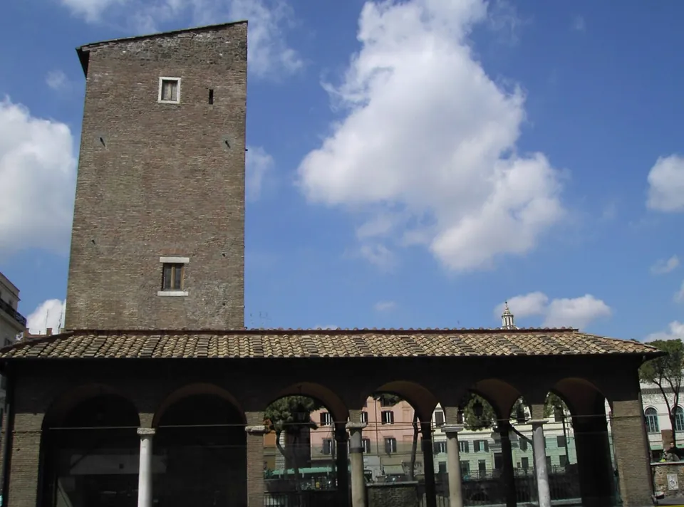 A tall, plain medieval brick tower rising behind a tiled-roof arcade of round arches on slender columns, under a blue sky.