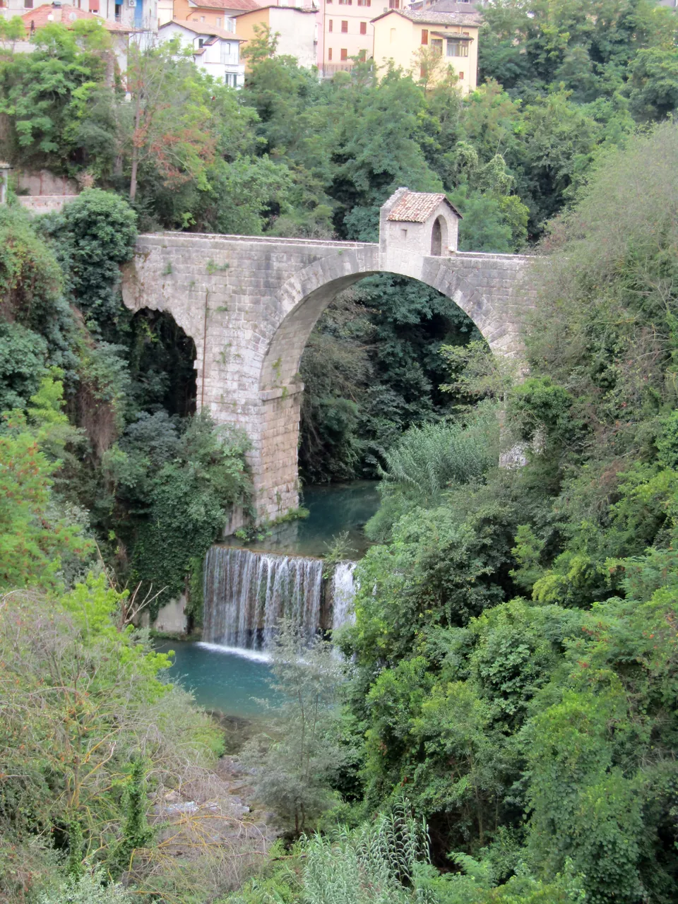 A two-arched stone bridge with a small house-like structure at its centre spans a green ravine, with a waterfall dropping into a pool below and houses at the top of the frame.