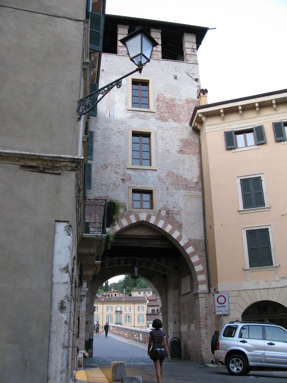A tall tower of patched plaster and brick over a round archway edged with alternating pale stone and brick; through the arch a cobbled deck with a low parapet leads away towards houses on the far bank. A street lamp hangs from the wall on the left and a car is parked on the right.