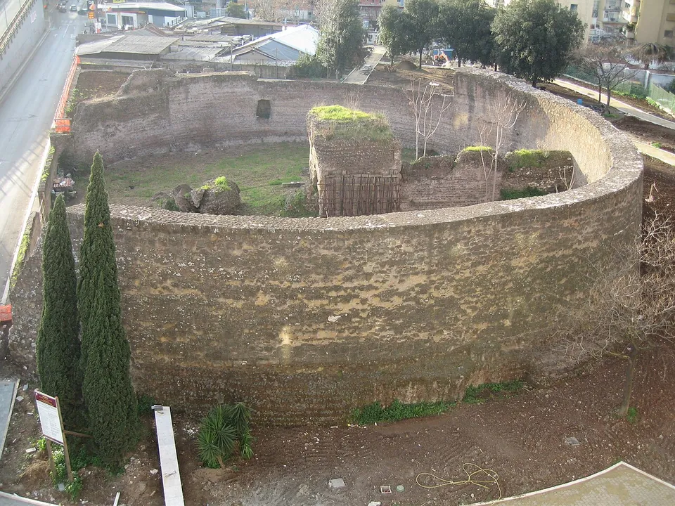 The tomb seen from above, a thick curved wall of rough masonry enclosing a lawn, with a square block of masonry in the middle of the ring, cypress trees beside the wall and a road on the left.