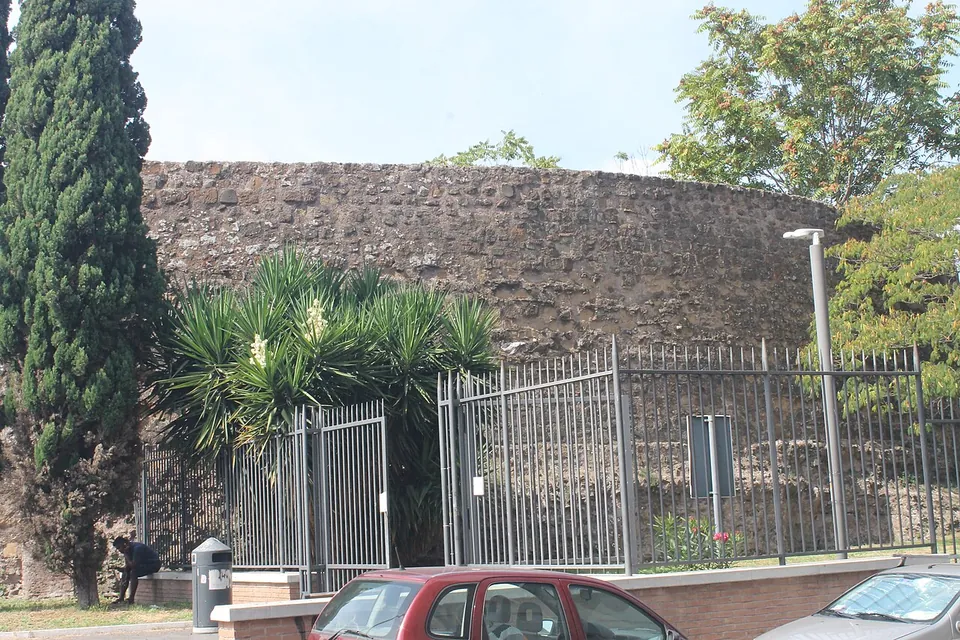The curved wall of the tomb rising behind a metal railing with a gate, a cypress and palms in front, and cars parked along the kerb.