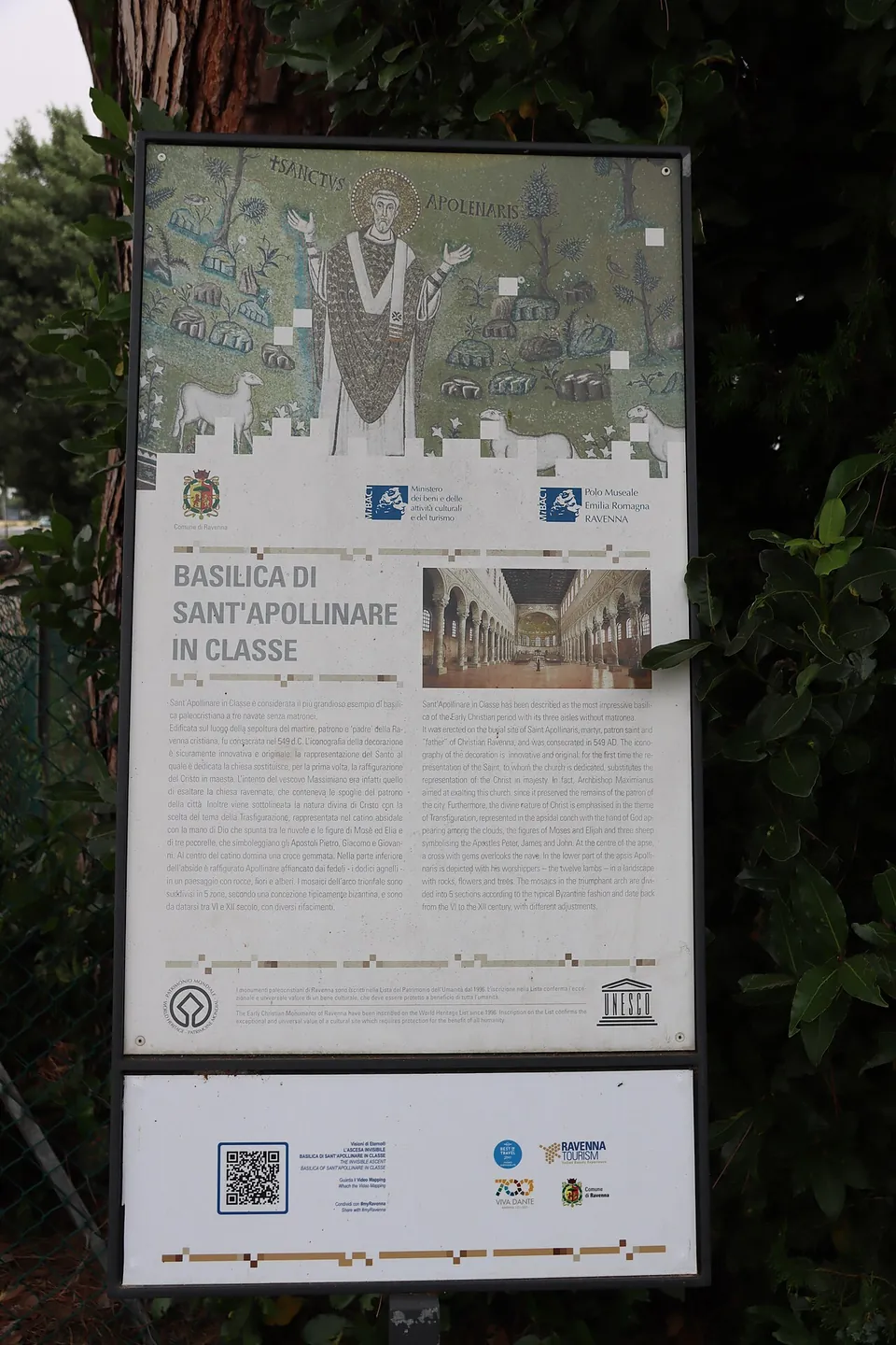 A tall information board under trees, headed "Basilica di Sant'Apollinare in Classe", with a picture of the mosaic saint at the top, a photograph of the nave and text in Italian and English, and the logos of the Ministry of Culture, the regional museum service and UNESCO.