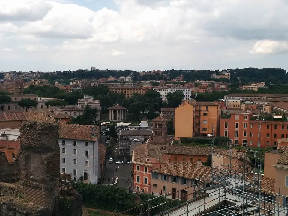 A high view over the red and ochre roofs of Rome under a cloudy sky, with a small round colonnaded temple among trees in the middle distance and green hills on the horizon; ancient brick walls and scaffolding in the foreground.