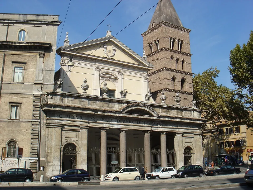 A church front on a busy street, with a portico of four columns under a balustrade and a pale pedimented façade above, beside a tall brick bell tower with rows of arched openings and a pointed spire; cars are parked along the kerb.